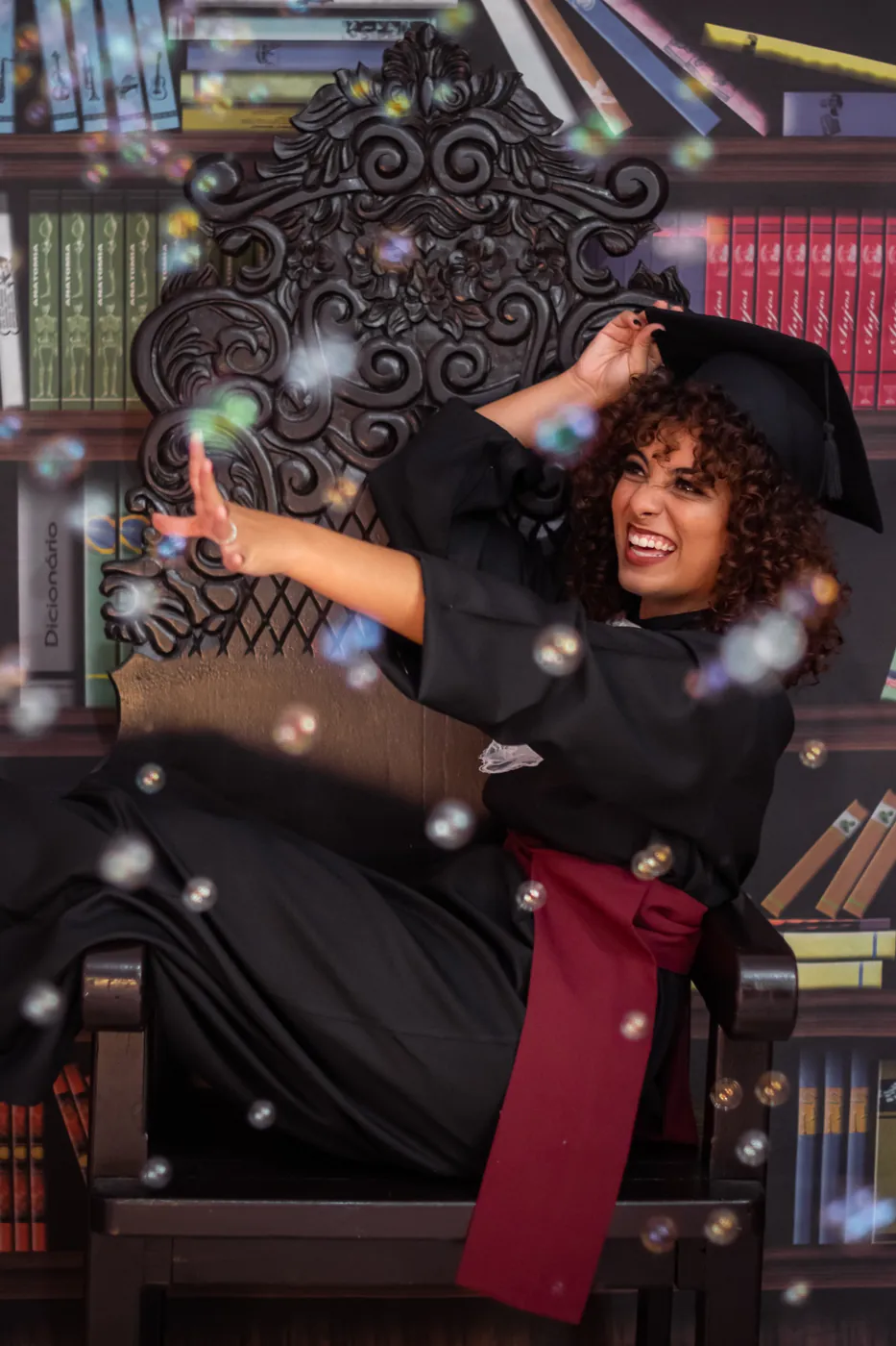 Formanda rindo em cadeira de biblioteca com bolhas de sabão no ar, ensaio externo