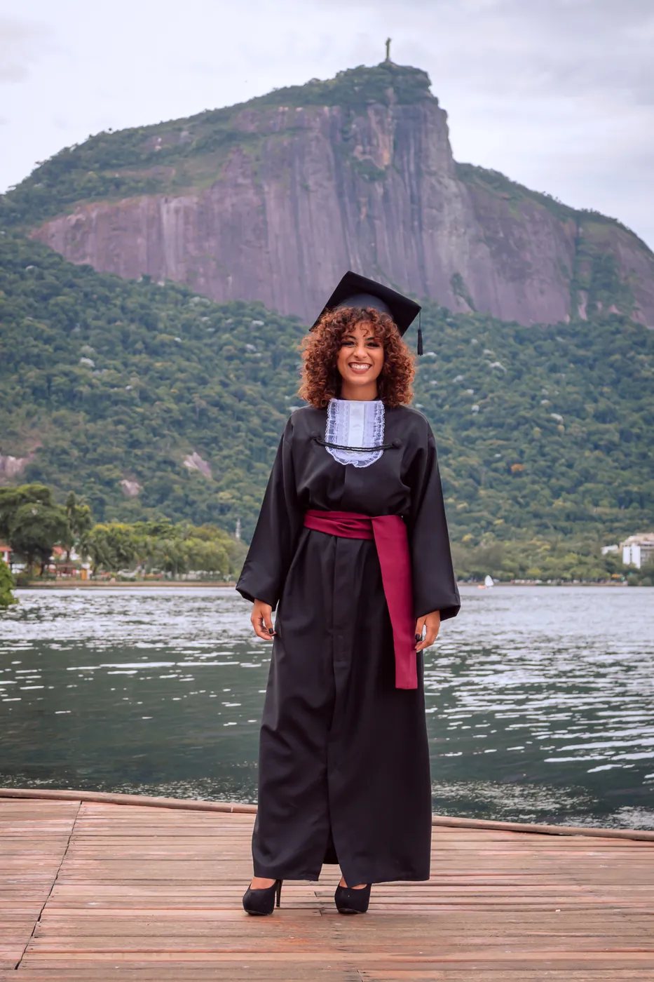 Formanda de beca e capelo sorrindo em pier de madeira com montanhas ao fundo, ensaio de formatura em locação externa